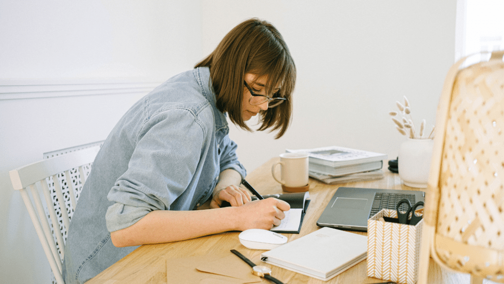 Woman writing in a notebook in frot of a laptop